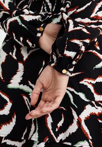 Black fabric dress with a bold red, white, and green abstract pattern. Features gathered sleeves, button accents, and a flowing design.