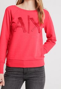 Sudadera color coral con un escote amplio, mangas raglán, logo “AIX” texturizado y dobladillo de canalé. Combinada con jeans gris oscuro.