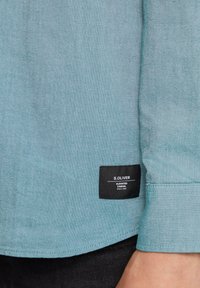 Lichtblauw, geweven stoffen shirt met een label waarop het "S.OLIVER" logo staat, een zwart rechthoekig label en gestikte manchetdetails.