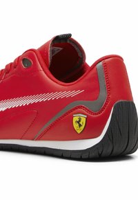 Sneaker rosso a scarpa bassa con suola nera, striscia diagonale bianca e logo giallo Ferrari sul lato vicino al tallone.
