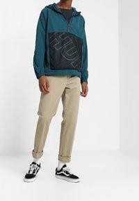 Tyrkysová větrovka s černou síťovou panely s logem, spárovaná s béžovými vyhrnutými kalhotami a černými teniskami.