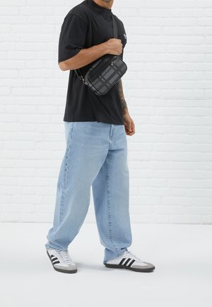 Man in een zwart t-shirt en lichtblauwe jeans, met een kleine zwarte schoudertas met een geruit patroon, staand tegen een witte bakstenen muur.