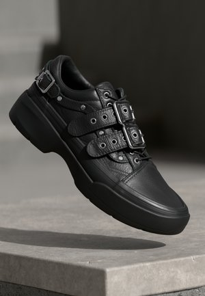 Schwarzer Ledersneaker mit doppelten Schnallenriemen, Schnürung vorne und dicker Gummisohle, der auf einer erhöhten Steinfläche vor dunklem Hintergrund ruht.