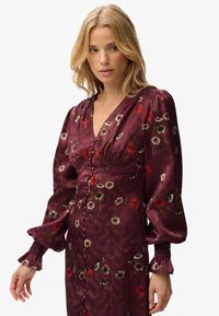 Vestido floral malva con escote en V profundo, mangas abullonadas y cierre de botones en la parte delantera. Presenta patrones de flores rojas y blancas sobre una tela suave.