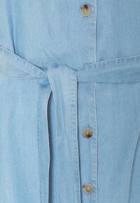 Ανοιχτό μπλε τζιν πουκάμισο με κουμπιά μπροστά, ζώνη στη μέση και ασημί κουμπιά κατά μήκος της παλιάς. Μαλακή υφή, ανεπίσημος σχεδιασμός.