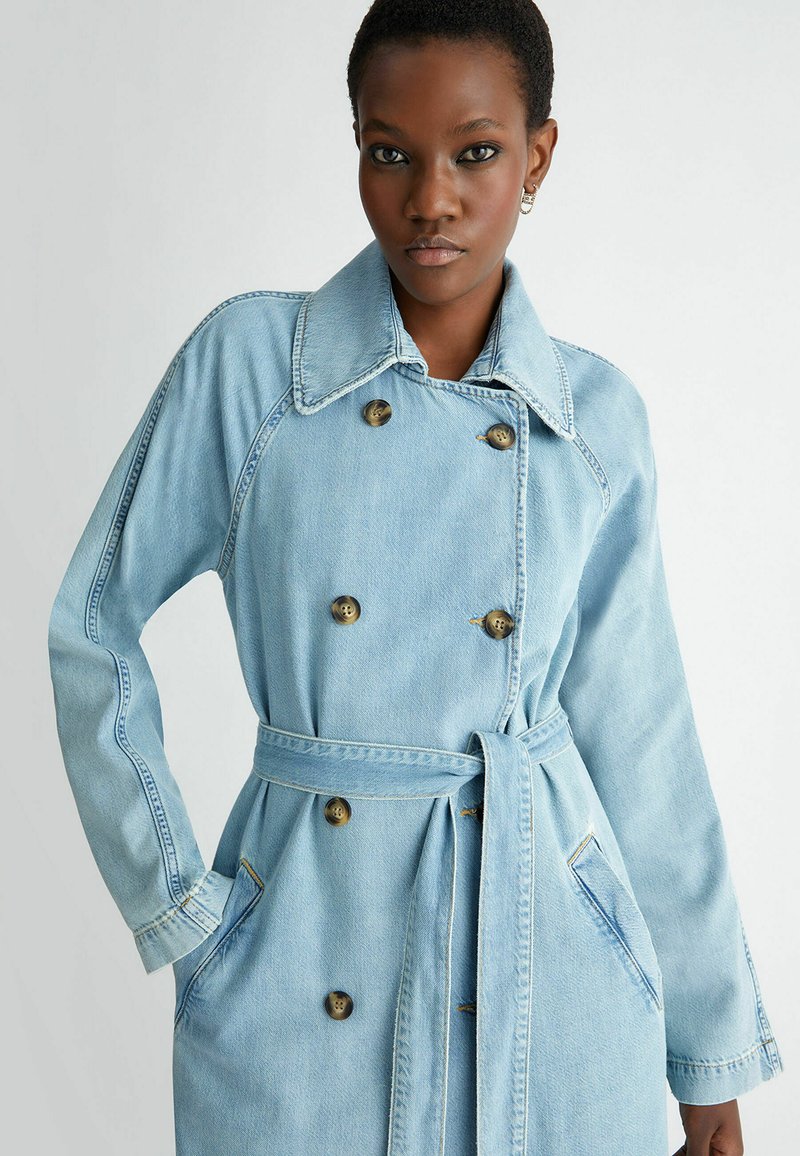Cappotto doppiopetto in denim azzurro chiaro con ampio colletto, bottoni in ottone, cintura e tasche laterali. Presenta cuciture a contrasto e una vestibilità rilassata.