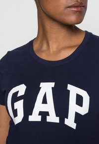 Marinblå kortärmad t-shirt i bomull, med stora vita bokstäver "GAP" över bröstet. Klassisk rundhalsad design.