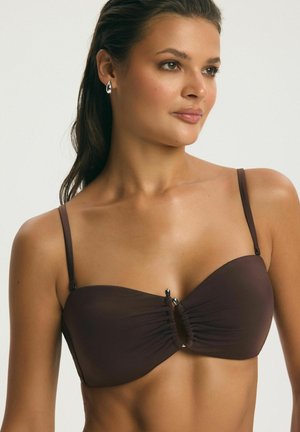 Žena nosi tmavohnedý bikiny top bez ramienok s tenkými ramienkami na pleciach a striebornými náušnicami, pozerá sa doprava na jednoduchom pozadí.