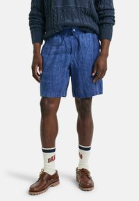 Pantaloni corti in lino di colore blu navy con una vestibilità comoda, vita elastica e cordoncino. Indossati con scarpe da barca marroni e calze bianche con scritte rosse.