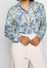 Long-sleeve, button-up shirt in a blue, green, and brown abstract pattern. Collared with buttoned cuffs, paired with cream-colored trousers.
