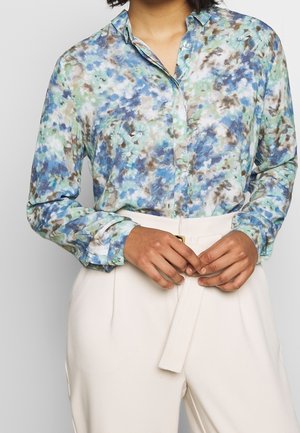 Long-sleeve, button-up shirt in a blue, green, and brown abstract pattern. Collared with buttoned cuffs, paired with cream-colored trousers.