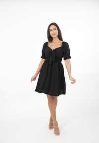 Schwarzes Kleid mit Puffärmeln und einer Bindefront. Verfügt über ein gerafftes Oberteil und einen fließenden Rock. Das Model trägt Sandalen mit transparenten Absätzen.
