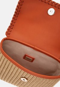 Kudotusta olkakassista, jossa on vaaleanruskea nahkasisustus, napsulukko ja kohokuvioitu brändin etiketti. Yksityiskohtana on punotun reunan viimeistely.