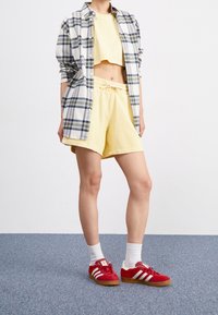 Person klädd i en gul kortärmad topp och shorts, lager på lager med en blå och vit rutig skjorta, vita strumpor och röda Adidas-sneakers på en blå matta.