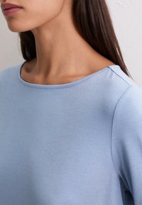 Kobieta w jasnoniebieskiej koszulce z okrągłym dekoltem, odsłaniająca szyję, ramię i część twarzy, z długimi ciemnymi włosami.