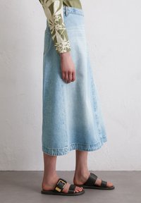 Light blue denim midi skirt with a flared shape, raw hem, and faded texture, paired with black sandals featuring a gold buckle detail.