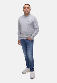 Maglietta grigia a maniche lunghe con un'illustrazione floreale e testo sul petto, abbinata a jeans blu e sneakers grigio chiaro.