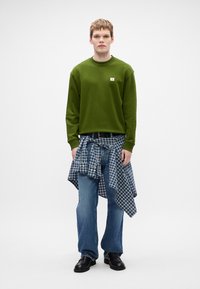 Sweatshirt verde com um pequeno logotipo, camisa de xadrez azul presa à cintura, jeans azul claro e sapatos pretos. Design simples e ajuste descontraído.