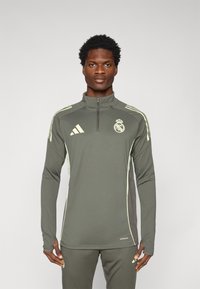 Dugi rukav sportski top u maslinastoj boji s četvrtim patent zatvaračem, kontrastnim svijetlozelenim bočnim panelima i logom Real Madrida na prsima.
