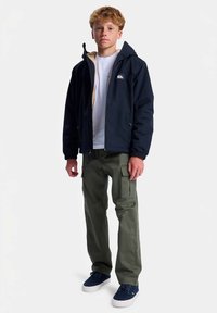 Băiat adolescent în picioare, îmbrăcat cu o jachetă cu glugă de culoare bleumarin, un tricou alb, pantaloni cargo oliva și adidași bleumarin, pe un fundal alb simplu.