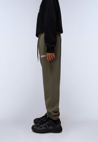 Olivgrüne Jogginghose mit elastischem Bund, gerippten Beinen und seitlichen Taschen, kombiniert mit schwarzen Sportschuhen mit strukturierten Sohlen.