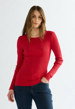 Giovane donna con lunghi capelli castano chiaro che indossa una maglia rossa a costine aderente a maniche lunghe e jeans blu scuro, in piedi con una mano in tasca.