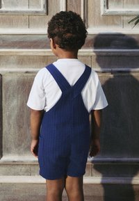 Blue ribbed overalls with adjustable straps, worn over a white t-shirt, viewed from behind against a wooden background.