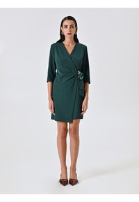 Robe portefeuille verte avec des manches trois-quarts, un détail de ceinture à nouer et une accent doré. Tissu lisse ; design jusqu'au genou. Portée avec des escarpins pointus bordeaux.