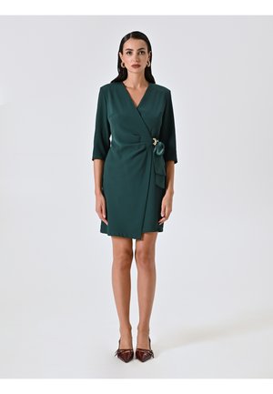 Grünes Wickelkleid mit Dreiviertelärmeln, einem taillierten Gürtel und einem goldenen Akzent. Glatte Stoffqualität; knielanges Design. Tragen Sie es mit spitzen bordeauxfarbenen Schuhen.