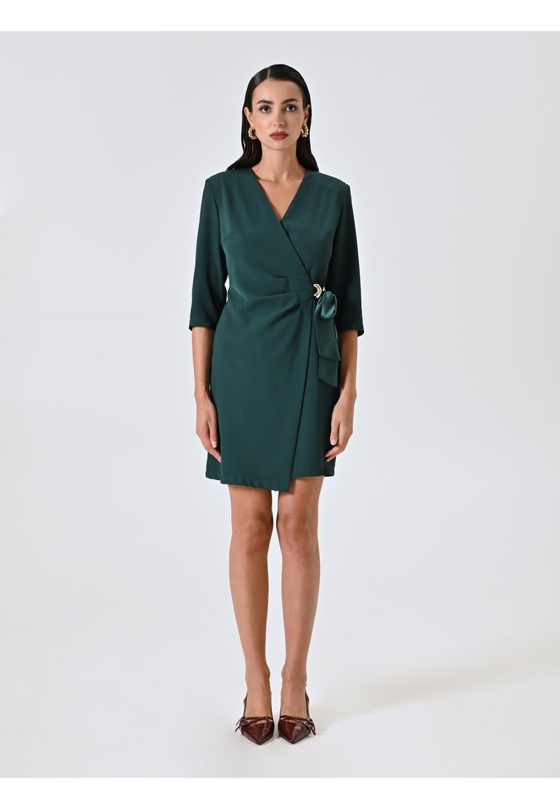 Robe portefeuille verte avec des manches trois-quarts, un détail de ceinture à nouer et une accent doré. Tissu lisse ; design jusqu'au genou. Portée avec des escarpins pointus bordeaux.