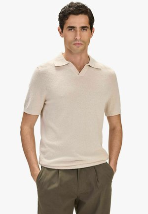 Man met donker haar, gekleed in een beige overhem met korte mouwen en een kraag, en olijfgroene broek, staand met handen in de zakken tegen een witte achtergrond.