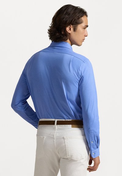 Camisa azul de manga longa com colarinho e punhos abotoados, combinada com jeans brancos. A camisa tem uma textura suave e um design ajustado.