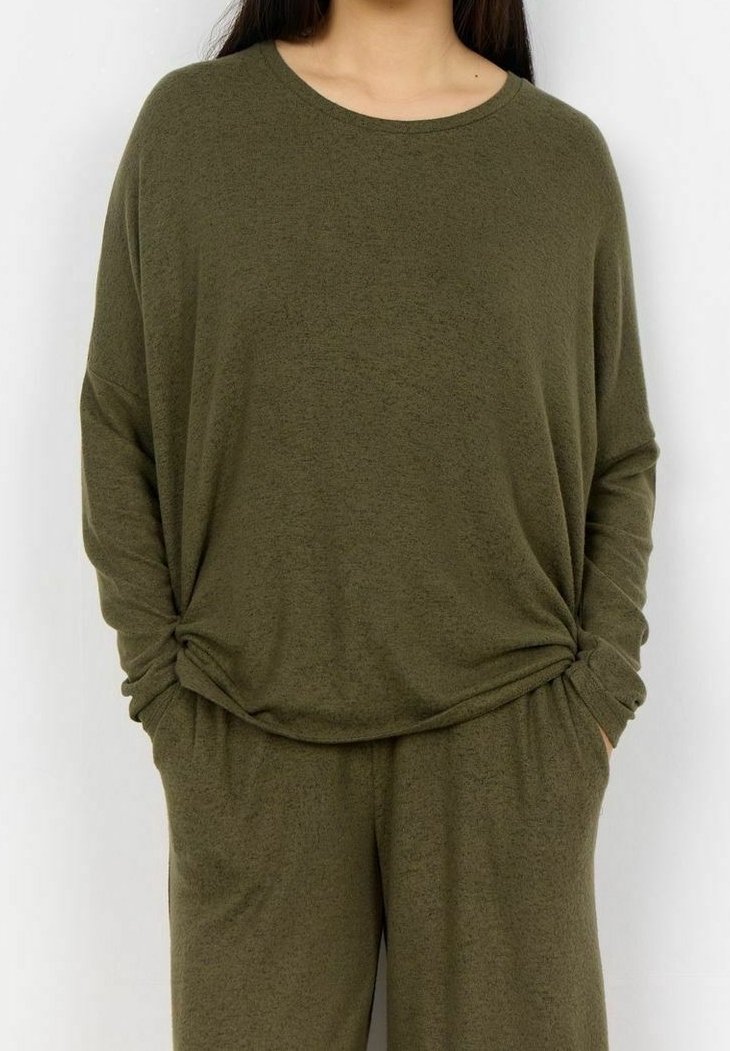 Frau trägt ein locker sitzendes, olivgrünes Langarmoberteil und passende, weit geschnittene Hose mit Händen in den Taschen vor einem einfarbigen Hintergrund.