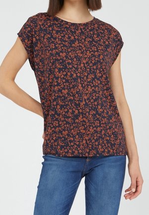 Kobieta w granatowej bluzce z krótkim rękawem z małym pomarańczowym kwiatowym wzorem i niebieskich dżinsach, z jedną ręką opartą na biodrze.