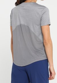 Camiseta deportiva gris con cuello redondo, mangas cortas y un panel trasero de malla para mayor transpirabilidad, con un logo de marca sutil.