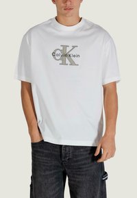 Biała bawełniana koszulka z dużym szaro-czarnym logo "Calvin Klein", krótkimi rękawami i okrągłym dekoltem. Połączona z ciemnymi dżinsami.