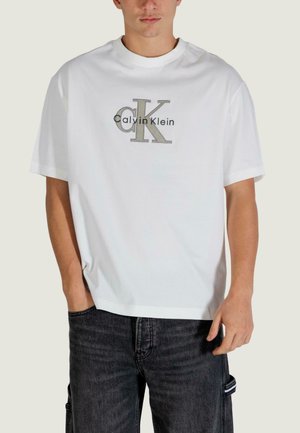 Biała bawełniana koszulka z dużym szaro-czarnym logo "Calvin Klein", krótkimi rękawami i okrągłym dekoltem. Połączona z ciemnymi dżinsami.