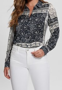 Blouse à manches longues bleu marine et crème avec motif paisley et accents rayés, col boutonné, associée à un jean blanc.