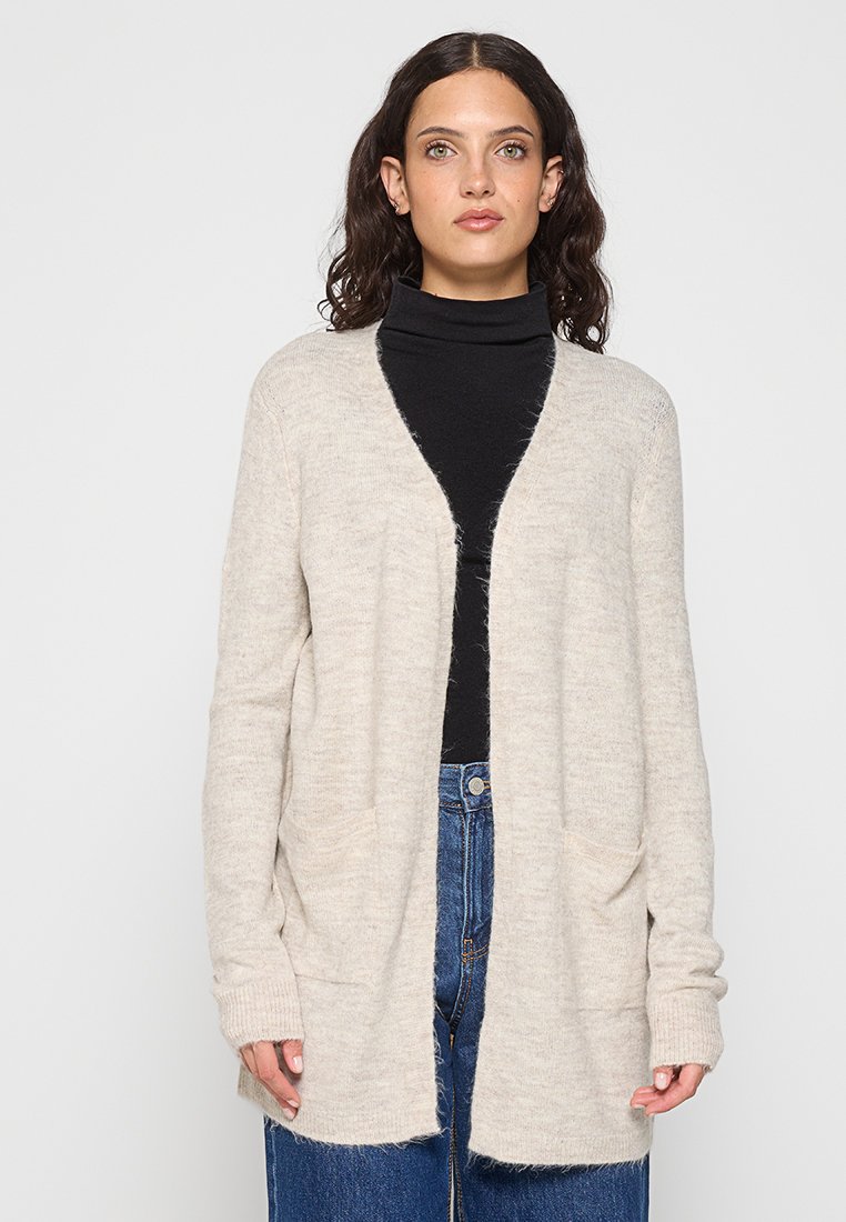 Beige gebreide cardigan met open voorkant, lange mouwen en zijzakken. Gelaagd stoffen met een zachte afwerking. Draag over een zwarte coltrui.