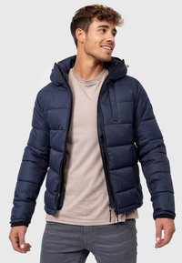Mørkeblå dunjakke med hætte, der har et quiltet design, lynlås foran og sidelommer. Bæret over en lys beige skjorte.