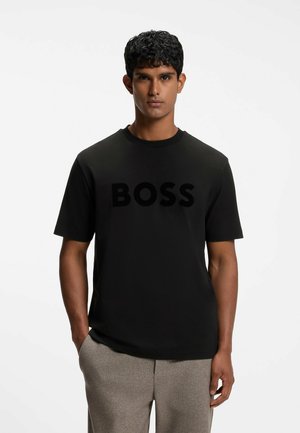 Mand med mørkt krøllet hår iført en sort T-shirt med "BOSS" print og lysegrå bukser, stående med hænderne i lommerne mod hvid baggrund.