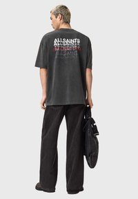 Sort oversized t-shirt med gentaget "ALLSAINTS" grafik i grå og rød. Kombineret med sorte bukser med vide ben. Modellen holder en sort taske.