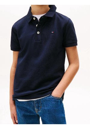 Garçon portant un polo bleu marine à manches courtes avec trois boutons blancs et un jean bleu, une main dans la poche, sur fond blanc.
