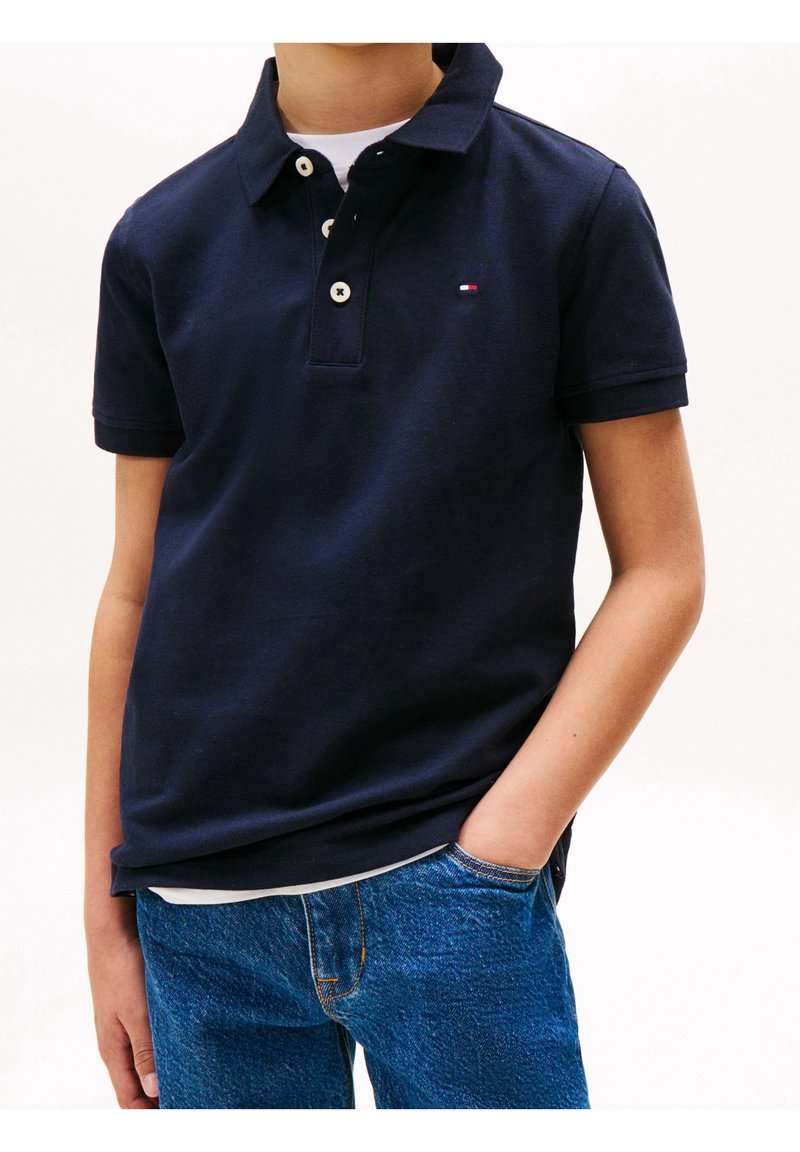 Garçon portant un polo bleu marine à manches courtes avec trois boutons blancs et un jean bleu, une main dans la poche, sur fond blanc.