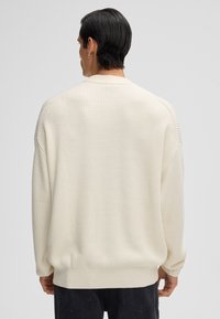 Mann mit kurzem dunklem Haar, der einen cremefarbenen gerippten Pullover trägt, steht mit dem Rücken zu einem schlichten hellen Hintergrund.