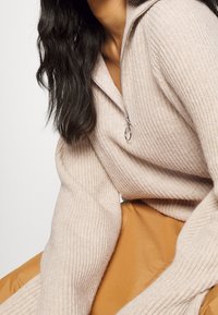 Hellrosa gerippter Strickpullover mit Reißverschluss am Kragen und langen Ärmeln, kombiniert mit einem glatten, gedämpften orangefarbenen Stoff darunter.