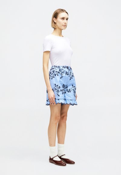 Γυναίκα που στέκεται φορώντας λευκό εφαρμοστό t-shirt, μπλε σορτς με κεντήματα από λουλούδια και βολάν στο τελείωμα, λευκές κάλτσες και σκούρα καφέ παπούτσια Mary Jane.