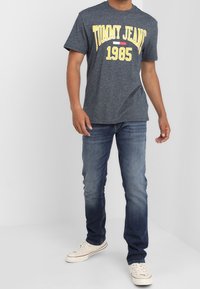 Camiseta gris oscuro con el texto amarillo "TOMMY JEANS 1985", combinada con jeans de mezclilla azules y zapatillas blancas, de pie contra un fondo claro.