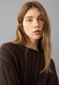 Brauner Strickpullover mit gerippter Struktur, weitem Ausschnitt und lockerem Sitz. Das Modell hat langes, welliges Haar und einen neutralen Hintergrund.