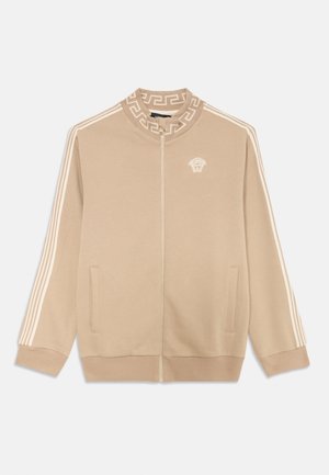 Chaqueta beige con cremallera y cuello alto, mangas a rayas blancas y un patrón de llaves griegas. Incluye bolsillos frontales y un logotipo bordado.